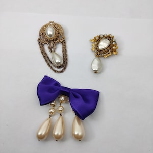 Peut inclure: Trois broches dorées avec des accents de perles. Une broche présente une grosse perle avec une chaîne dorée et une petite perle pendante. Une autre broche présente une grosse perle avec un motif floral doré. La troisième broche présente un nœud en satin violet avec trois gouttes de perles.