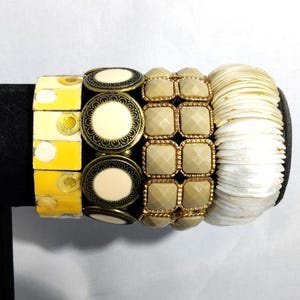 May include: A collection of bracelets in various styles. One bracelet features yellow rectangular beads, another has cream-colored circular beads with a black and gold border, and a third has square, tan beads. The last bracelet is made of white shell-like pieces.