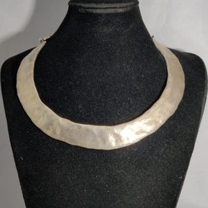 May include: A silver-toned collar necklace with a hammered texture, displayed on a black mannequin. The necklace has a wide, curved design that sits around the neck. The metal has a slightly uneven surface.