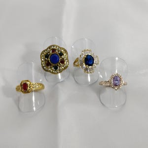 May include: Four rings with colorful gemstones are displayed. The rings feature gold and silver bands with red, blue, and purple stones. One ring has a large blue stone surrounded by smaller white stones.