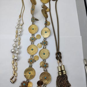 Peut inclure: Trois colliers en or de styles différents. Un collier présente une chaîne avec de grandes pièces d'or ornées d'accents turquoise. Un autre collier a une chaîne avec de petites perles dorées et un brin de perles. Le troisième collier a une chaîne avec un pompon.