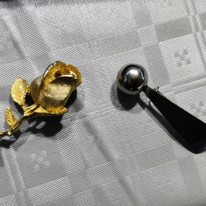 May include: A gold rose brooch with a stem and leaves, and a silver and black brooch with a round ball on top.