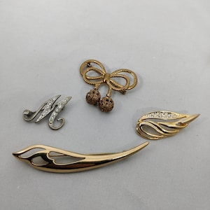 Może przedstawiać: Cztery złote broszki z cyrkoniami. Jedna broszka to litera M, jedna to kokarda z dwoma zwisającymi kulkami, a dwie są w kształcie liścia.
