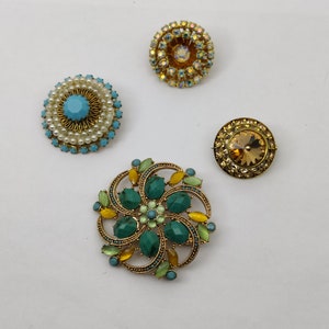 Può includere: Quattro spille vintage con strass e perle colorati. Le spille sono dorate e presentano una varietà di design, tra cui un motivo floreale, un design circolare con perle e un design circolare con strass.