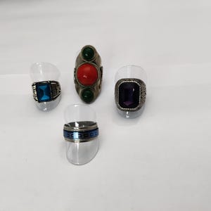 Peut inclure: Quatre bagues en argent avec des pierres précieuses colorées. Les bagues sont exposées sur de petits supports en plastique transparent. Une bague a une pierre rouge, verte et verte. Une autre bague a une pierre bleue. Une autre bague a une pierre violette. La dernière bague a une bande bleue et argentée.
