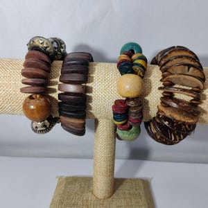 May include: Five wooden bracelets on a display stand. The bracelets are made of different types of wood and have various designs, including round beads, flat pieces of wood, and coconut shells.