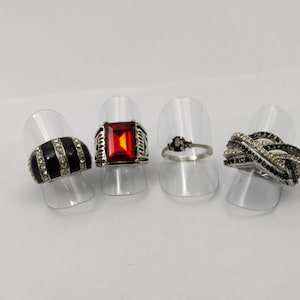 Peut inclure: Quatre bagues en argent avec des pierres noires et blanches sur des supports d'exposition. Les bagues présentent différents styles, dont une bague à pierre rouge, une bague à pierre noire et deux bagues à pierres noires et blanches.