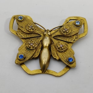 Pode incluir: Um broche de borboleta em tom dourado com strass azuis e rosa. A borboleta tem asas detalhadas e um corpo texturizado.