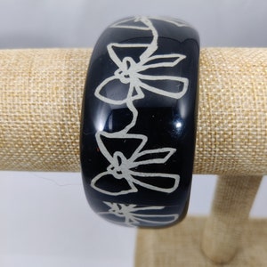 May include: Black bangle bracelet with a white design of bows. The bangle is on a display stand.
