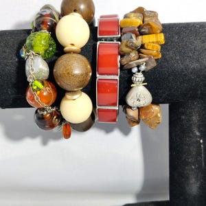 May include: A collection of beaded bracelets in various styles. One bracelet features large, round wooden beads in brown and cream colors. Another has rectangular red beads. A third bracelet is made of tiger eye stones.