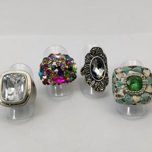 Könnte beinhalten: Vier Ringe mit unterschiedlichen Designs und Edelsteinen. Der erste Ring hat einen großen, klaren quadratischen Stein. Der zweite Ring hat einen Cluster aus bunten Steinen. Der dritte Ring hat einen schwarzen Stein in einer silbernen Fassung. Der vierte Ring hat einen grünen Stein in einer weißen und grünen Fassung.