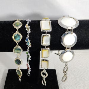 May include: Four bracelets displayed on a black velvet stand. The bracelets feature various designs, including abalone shell, colorful enamel, mother-of-pearl, and silver-tone metal. The bracelets are of different lengths and styles.