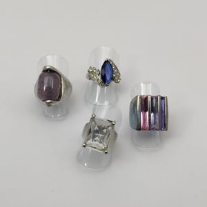 May include: Four silver rings with different colored gemstones. The rings are displayed on clear plastic ring holders. The rings feature a purple oval stone, a blue marquise stone, a clear rectangular stone, and a ring with multiple pink and purple rectangular stones.