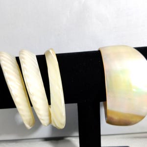 Könnte beinhalten: Vier elfenbeinfarbene Armreifen auf einem schwarzen Ständer. Drei haben ein geriffeltes Design, der vierte ist ein breiter Manschettenarmreif. Der Schmuck besteht aus hellem Material, möglicherweise Muschel oder Knochen.