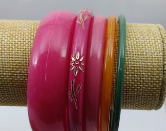 Può includere: Una collezione di braccialetti colorati nelle tonalità del rosa, arancione e verde. Il braccialetto più spesso è un fucsia vibrante, mentre altri sono rosa chiaro, arancione e un verde sottile. Un braccialetto rosa ha dettagli floreali dorati. I braccialetti sono esposti su una superficie beige.
