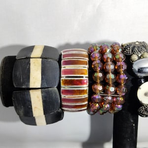 Peut inclure: Une collection de bracelets de différents styles. Le premier bracelet est noir avec une bande crème. Le second est un bracelet empilé et coloré avec des tons rouges et oranges. Le troisième est un bracelet de perles avec des perles irisées. Le dernier bracelet a une perle argentée et des éléments décoratifs.