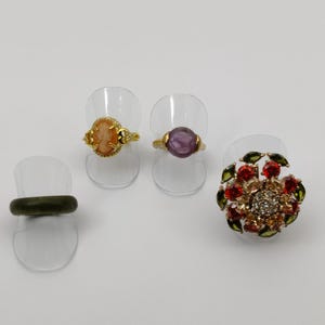 May include: Four rings on display stands. One ring has a cameo design with a gold setting. Another ring has a purple stone with a gold setting. A third ring has a cluster of red, green, and clear stones with a gold setting. The fourth ring is a simple green band.