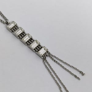 Puede incluir: Collar plateado con una serie de acentos rectangulares de cristal transparente. El diseño incluye una fila vertical de cristales, conectados por una cadena, con múltiples cadenas colgantes en la parte inferior. El collar está sobre un fondo blanco.