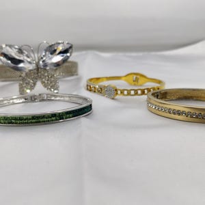 May include: An assortment of bracelets in various styles. One bracelet features a butterfly design with clear crystals. Other bracelets include a silver band with green stones, a gold chain link bracelet, and a gold band with clear crystals.