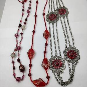 Puede incluir: Dos collares, uno con cuentas de vidrio rojo y transparente y el otro con cadenas plateadas y dijes de flores rojas.