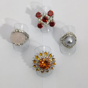 Op de afbeelding: Vier zilveren ringen met verschillende gekleurde edelstenen. De ringen worden gepresenteerd op kleine plastic ringhouders. De eerste ring heeft een grote rode edelsteen omgeven door kleinere heldere edelstenen. De tweede ring heeft een grote ovale roze edelsteen. De derde ring heeft een grote grijze edelsteen omgeven door kleinere heldere edelstenen. De vierde ring heeft een grote oranje edelsteen omgeven door kleinere heldere en gele edelstenen.