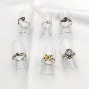 Puede incluir: Colección de anillos de plata exhibidos en soportes transparentes. Los anillos presentan varios diseños, incluyendo un nudo, una mariposa con detalles amarillos y piedras en púrpura, negro y azul. Fondo blanco.