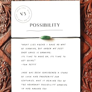 Puede incluir: Una pulsera de piedra de jade con un cordón negro, exhibida en una tarjeta blanca con la palabra "POSSIBILITY" y una cita de Tom Petty. La tarjeta también presenta un logotipo y el texto "VERSE THREE".