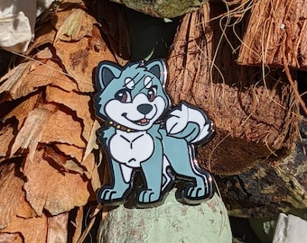 May include: A blue and white enamel pin of a cartoon dog with a white stripe across its face. The dog is wearing a gold collar and has a happy expression.