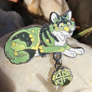 May include: A green and black wolf enamel pin with yellow flower accents, lying down. The pin has a dangling charm with a Celtic knot design. The pin is set on a white stone.