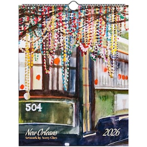 May include: A 2026 calendar featuring a watercolor painting of a New Orleans streetcar adorned with colorful beads. The artwork includes the number "504" and the text "New Orleans" and "Artwork by Avery Ches". The calendar is suspended by a metal ring.