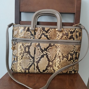 Op de afbeelding: Een handtas met slangenpatroon, een handvat aan de bovenkant en een afneembare schouderriem. De tas heeft een neutraal kleurenpalet van bruin, beige en grijs, met goudkleurige hardware-accenten. Het heeft een voorvak met ritssluiting en een gestructureerd ontwerp.
