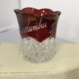 May include: A small, clear glass cup with a red, scalloped top. The cup has a cut glass design and the words "Columbus, O." are etched into the red top.