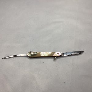May include: A small, vintage pocket knife with a mother-of-pearl handle and a silver blade. The knife has two blades, one pointed and one curved. The handle has a decorative design. The knife is open, showing both blades.