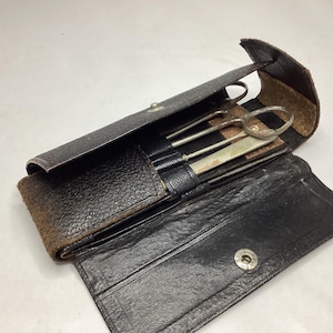 May include: An open, black leather case with a snap closure, revealing several silver medical instruments. The case has a textured exterior and a worn, brown edge. The instruments include a pair of scissors and several other tools.