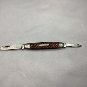 May include: A vintage pocket knife with two blades. The knife has a brown, textured handle and silver-colored metal bolsters. The blades are open, displaying their sharp edges. The knife is set against a plain, light gray background.
