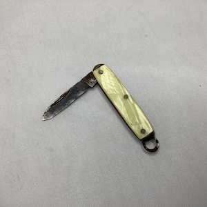 May include: A small, vintage pocket knife with a partially open blade. The handle is a light cream color with three small rivets. A small metal ring is attached to the end of the handle.