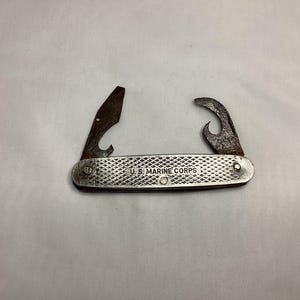 May include: Vintage U.S. Marine Corps pocket knife with a textured silver handle. The knife features a bottle opener and a can opener, both showing signs of age. The text "U.S. MARINE CORPS" is imprinted on the handle.
