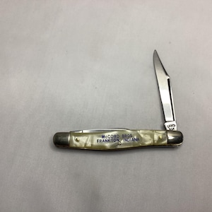 May include: A pocket knife with a partially open stainless steel blade. The handle is a pearlescent white and silver color. The text "McCORD BROS. FRANKTON, INDIANA" is printed on the handle.
