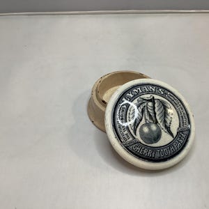 May include: A round, off-white ceramic container with a removable lid. The lid features a black and white illustration of a cherry with leaves and text that reads LYMAN'S CHERRY TOOTH PASTE, MONTREAL, CANADA, IMPARTS A DELIGHTFUL FRAGRANCE.