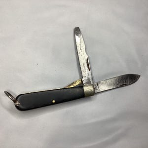 May include: A vintage pocket knife with a black handle and two open blades. The blades are silver and show signs of wear. A small metal ring is attached to the end of the handle.