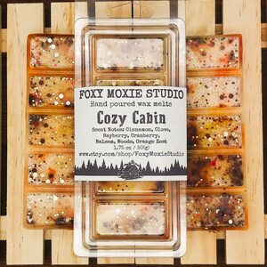 May include: Wax melts in a clear plastic container. The melts are a blend of orange, brown, and white with glitter. The label says "Foxy Moxie Studio" and "Cozy Cabin", with scent notes. The package is 50g.