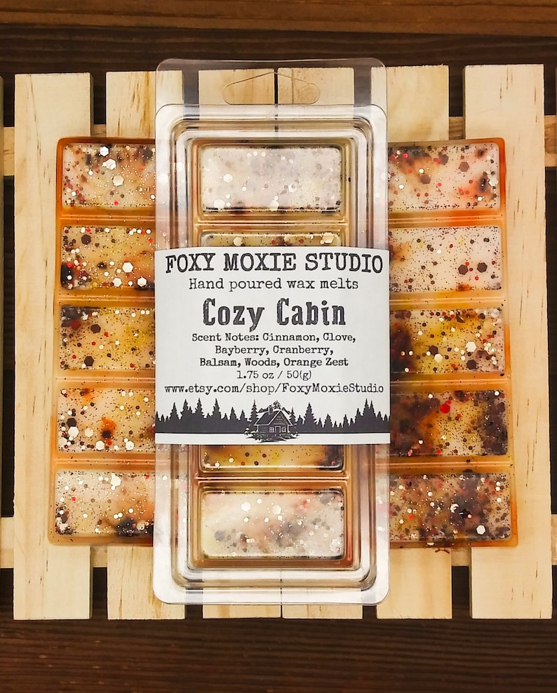 May include: Wax melts in a clear plastic container. The melts are a blend of orange, brown, and white with glitter. The label says "Foxy Moxie Studio" and "Cozy Cabin", with scent notes. The package is 50g.