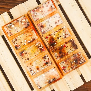 May include: Two rectangular wax melt bars in an orange hue, each divided into six sections. The wax is speckled with red, white, and gold glitter. The bars are set on a light wooden surface.