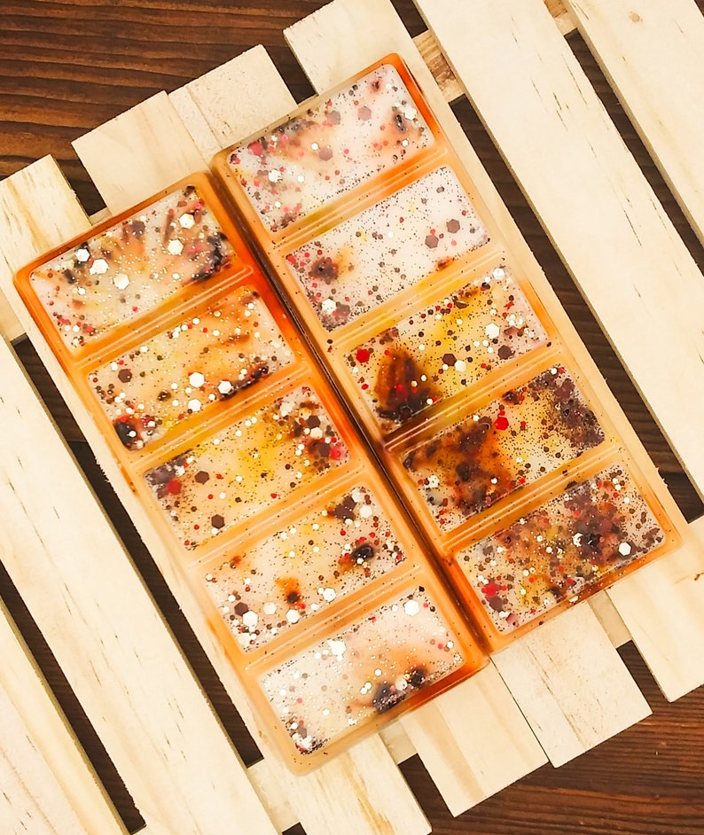 May include: Two rectangular wax melt bars in an orange hue, each divided into six sections. The wax is speckled with red, white, and gold glitter. The bars are set on a light wooden surface.