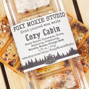 May include: A clear plastic container of hand-poured wax melts labelled "Cozy Cabin" by Foxy Moxie Studio. The label lists scent notes: Cinnamon, Clove, Bayberry, Cranberry, Balsam, Woods, and Orange Zest. The wax melts are 50g.