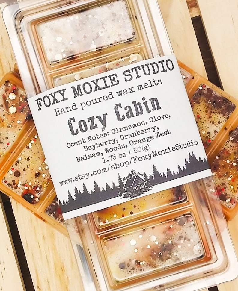 May include: A clear plastic container of hand-poured wax melts labelled "Cozy Cabin" by Foxy Moxie Studio. The label lists scent notes: Cinnamon, Clove, Bayberry, Cranberry, Balsam, Woods, and Orange Zest. The wax melts are 50g.