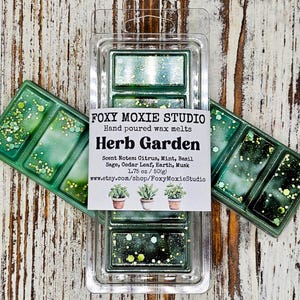 May include: Green wax melts in a clear plastic container. The label reads "Herb Garden" with scent notes and shop information. The wax melts are decorated with glitter.