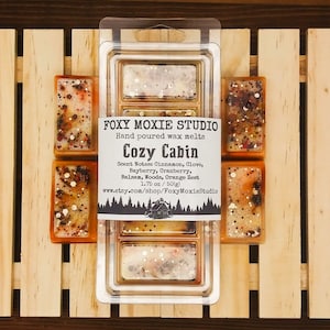 May include: A selection of hand-poured wax melts from Foxy Moxie Studio, labelled "Cozy Cabin." The wax melts are rectangular, presented in a clear plastic container. The scent notes include cinnamon, clove, bayberry, cranberry, balsam, woods, and orange zest. Individual wax melts are also visible.