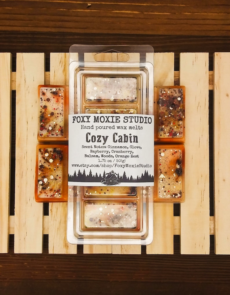 May include: A selection of hand-poured wax melts from Foxy Moxie Studio, labelled "Cozy Cabin." The wax melts are rectangular, presented in a clear plastic container. The scent notes include cinnamon, clove, bayberry, cranberry, balsam, woods, and orange zest. Individual wax melts are also visible.
