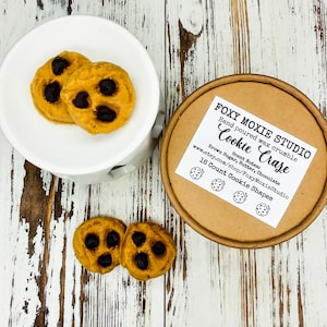 May include: A brown paper container with a white label that reads "FOXY MOXIE STUDIO Hand poured wax crumble Cookie Craze Brown Sugar, Butter, Chocolate Scent Notes www.etsy.com/shop/FoxyMoxieStudio 16 Count Cookie Shapes". The container is filled with 16 small, brown cookie-shaped wax melts.